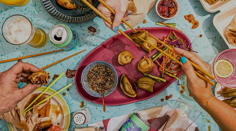 Overhead view of hands with chopsticks sharing a platter of dumplings, dipping sauce and drinks across a table